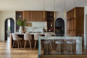 Modern kitchen with wood cabinets, glass panels, and white bar stools along a sleek countertop in natural lighting.