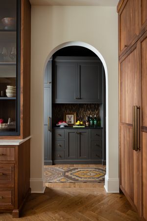 Arched doorway leads to a butler's pantry with dark gray cabinets and patterned tile floor beneath wood paneling.