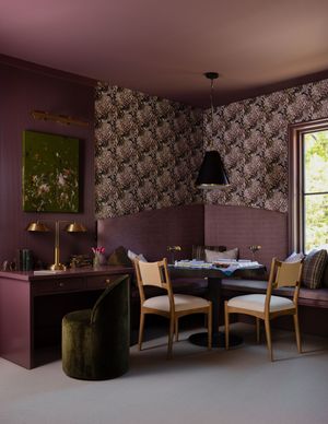 Moody dining nook with mauve walls, floral wallpaper, dark table, wooden chairs, green velvet ottoman, and brass lamp.