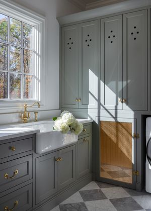 Elegant laundry room with sage green cabinets, brass hardware, farmhouse sink, white hydrangeas, and checkered marble floor.