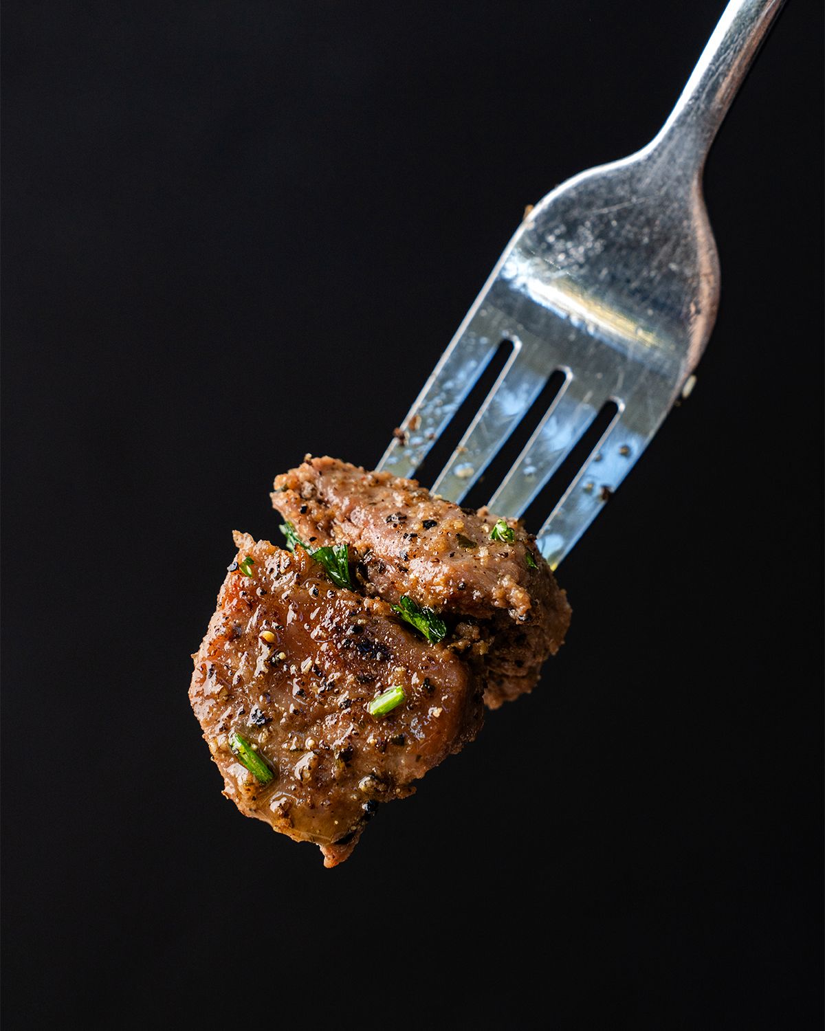 Well lit closeup image of seasoned and garnished bite size steak on a fork with a black background.