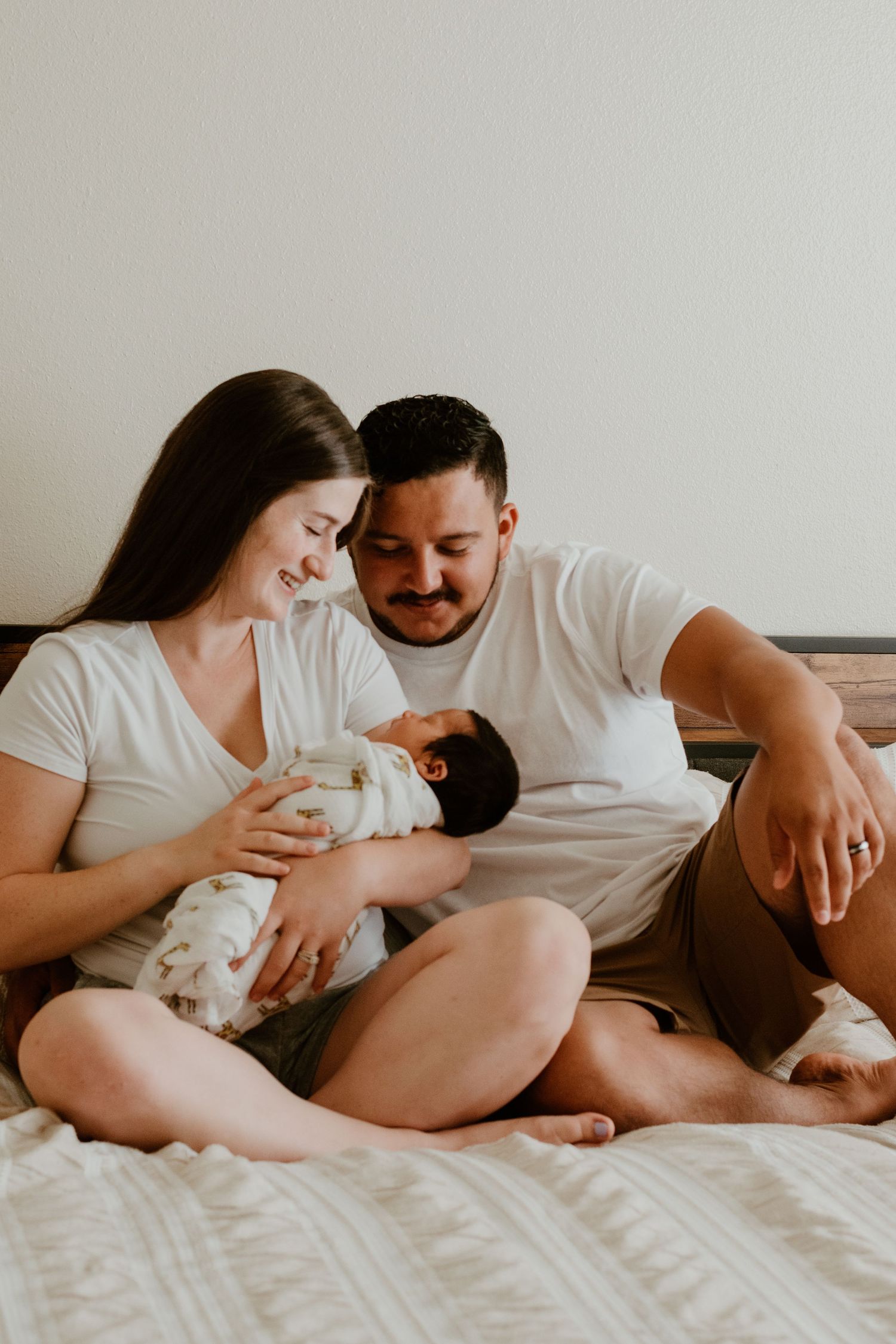 A couple in white shirts sits on a bed holding their newborn baby wrapped in a blanket.