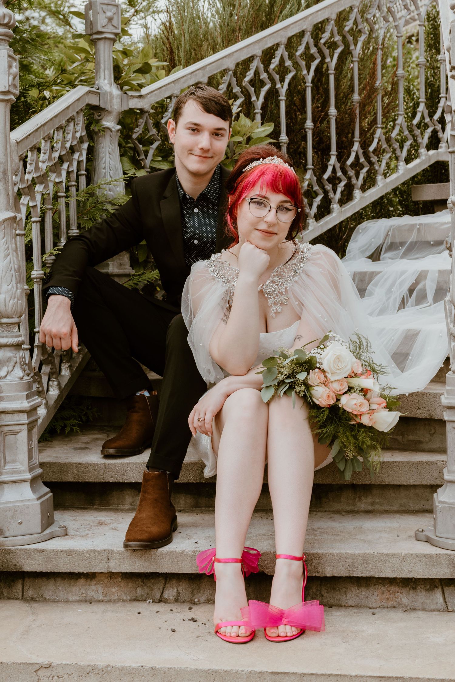 A couple sits on stone steps, the bride wearing a white dress with bright pink shoes and hair while holding a floral bouquet.