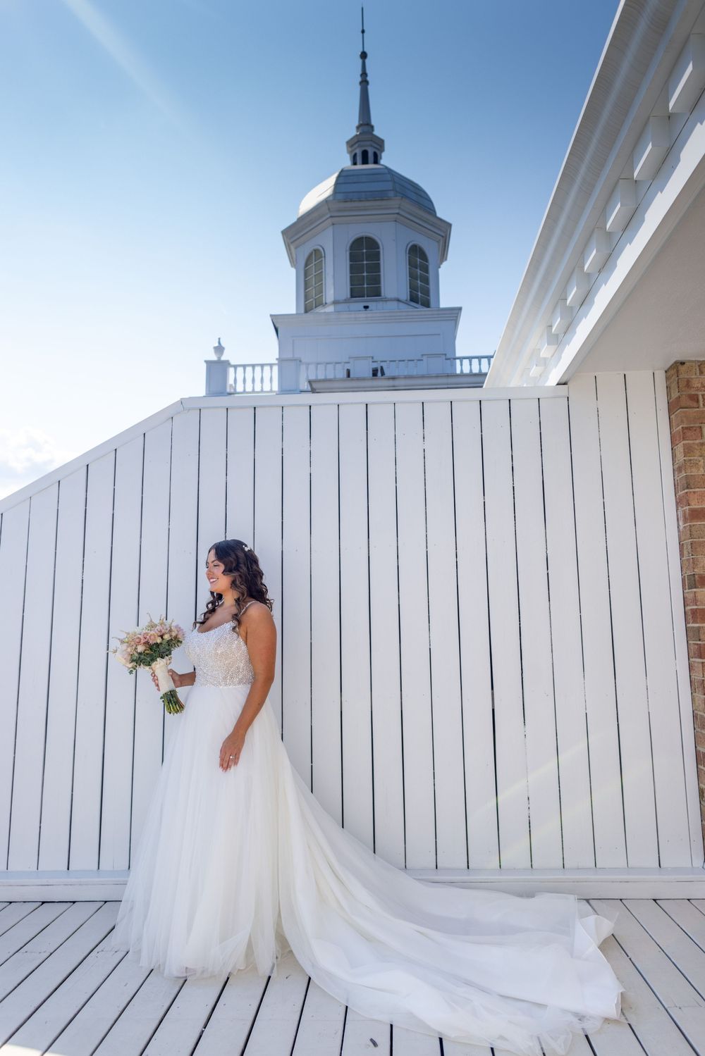 Elegant bride in a flowing white gown holding a bouquet beside a white wooden wall with a cupola tower.