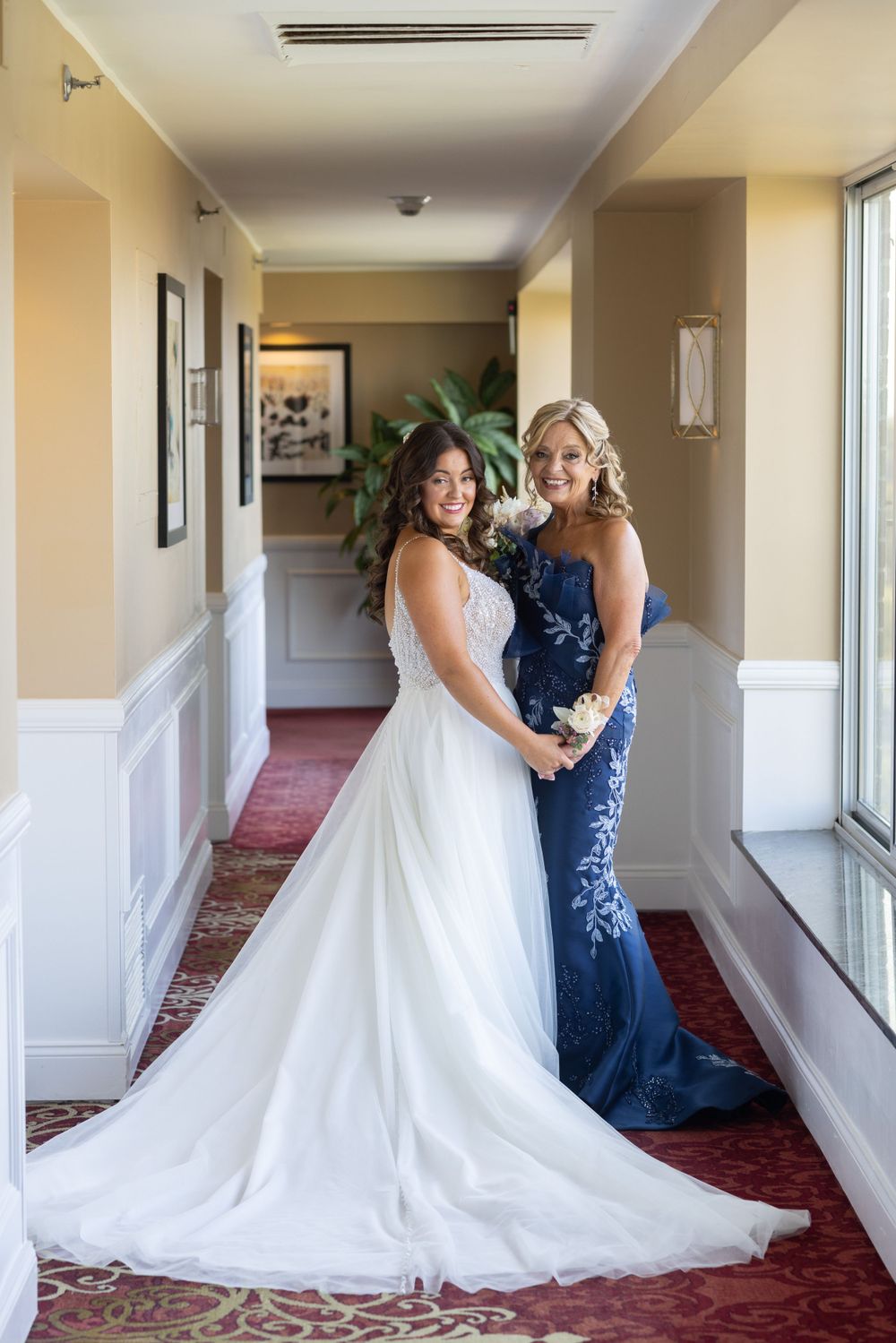 Bride in a white gown posing with a bridesmaid in a navy blue floral dress in a hotel hallway.