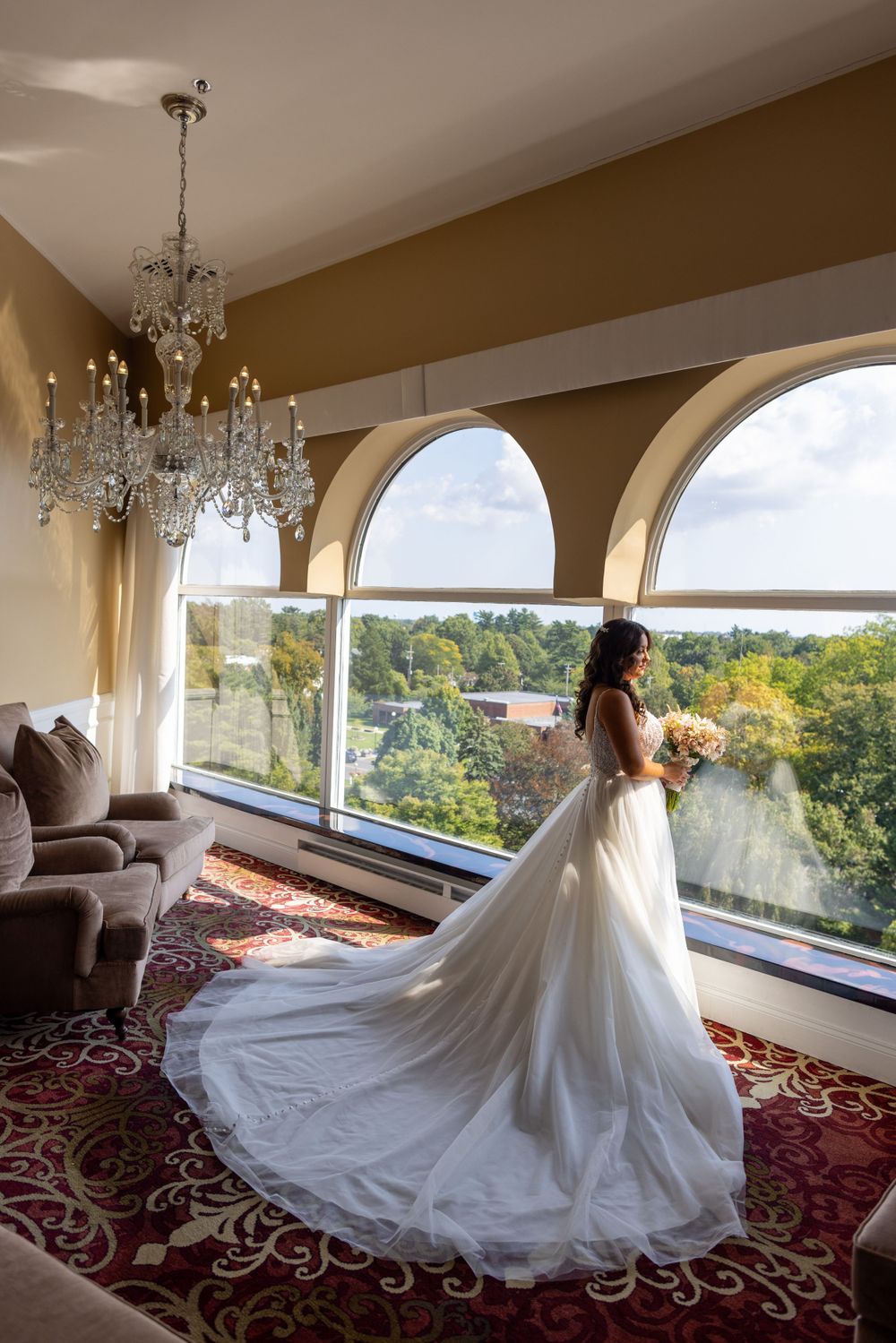 Bride in a long white gown holding a bouquet while standing before large arched windows with a scenic view.