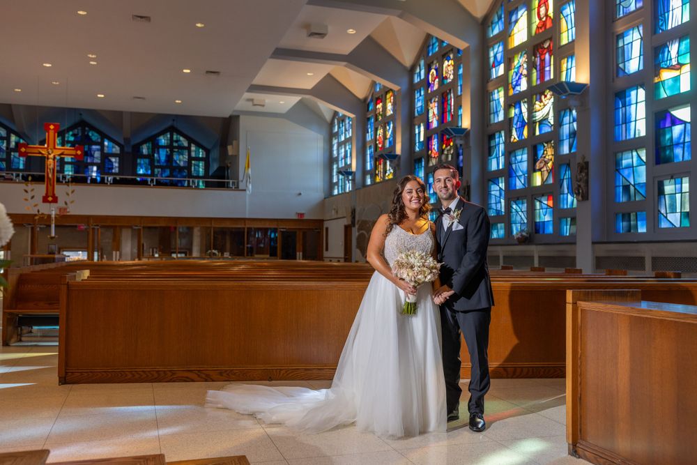 Bride and groom posing together inside a church with magnificent blue and multicolored stained glass windows behind them.