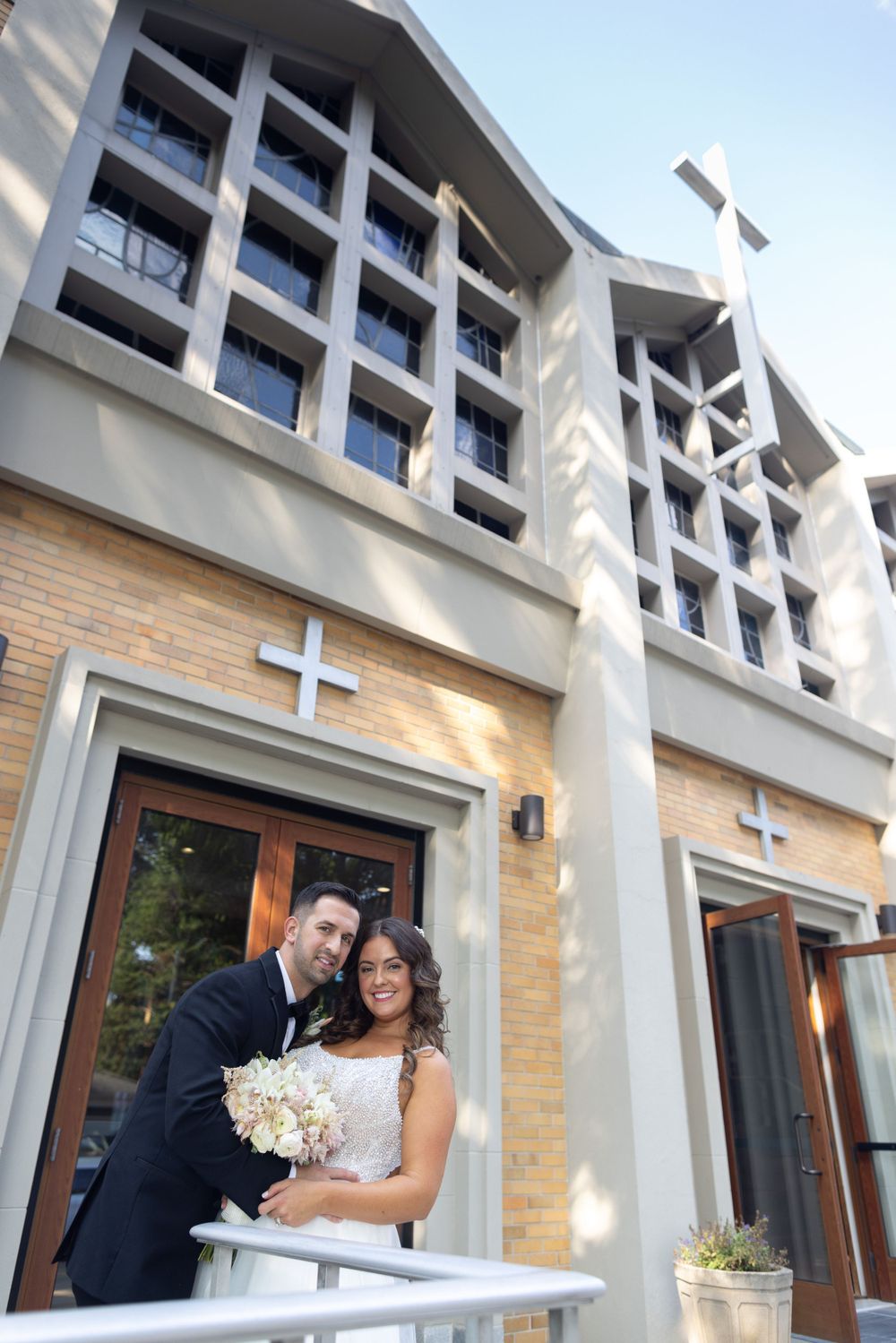 Bride and groom pose on church steps with crosses and brick architecture behind them on their wedding day.