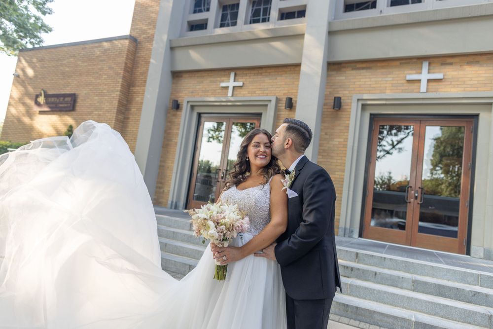 Bride's veil blows in wind as couple embraces outside brick church with crosses on wedding day.