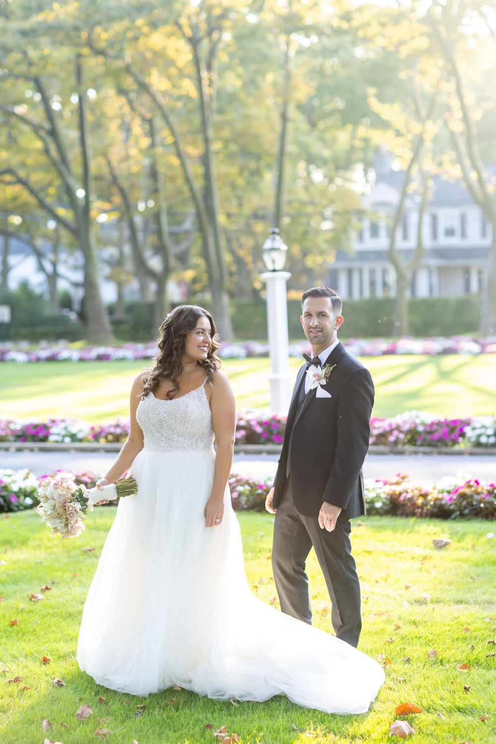 Bride in white gown and groom in black tuxedo walk through lush garden with colorful flowers at sunset.
