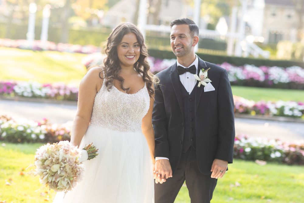 Smiling bride holding bouquet and groom in black tuxedo hold hands walking through flower garden.
