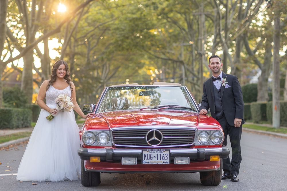 Bride and groom stand beside a classic red Mercedes convertible on a tree-lined road at sunset.