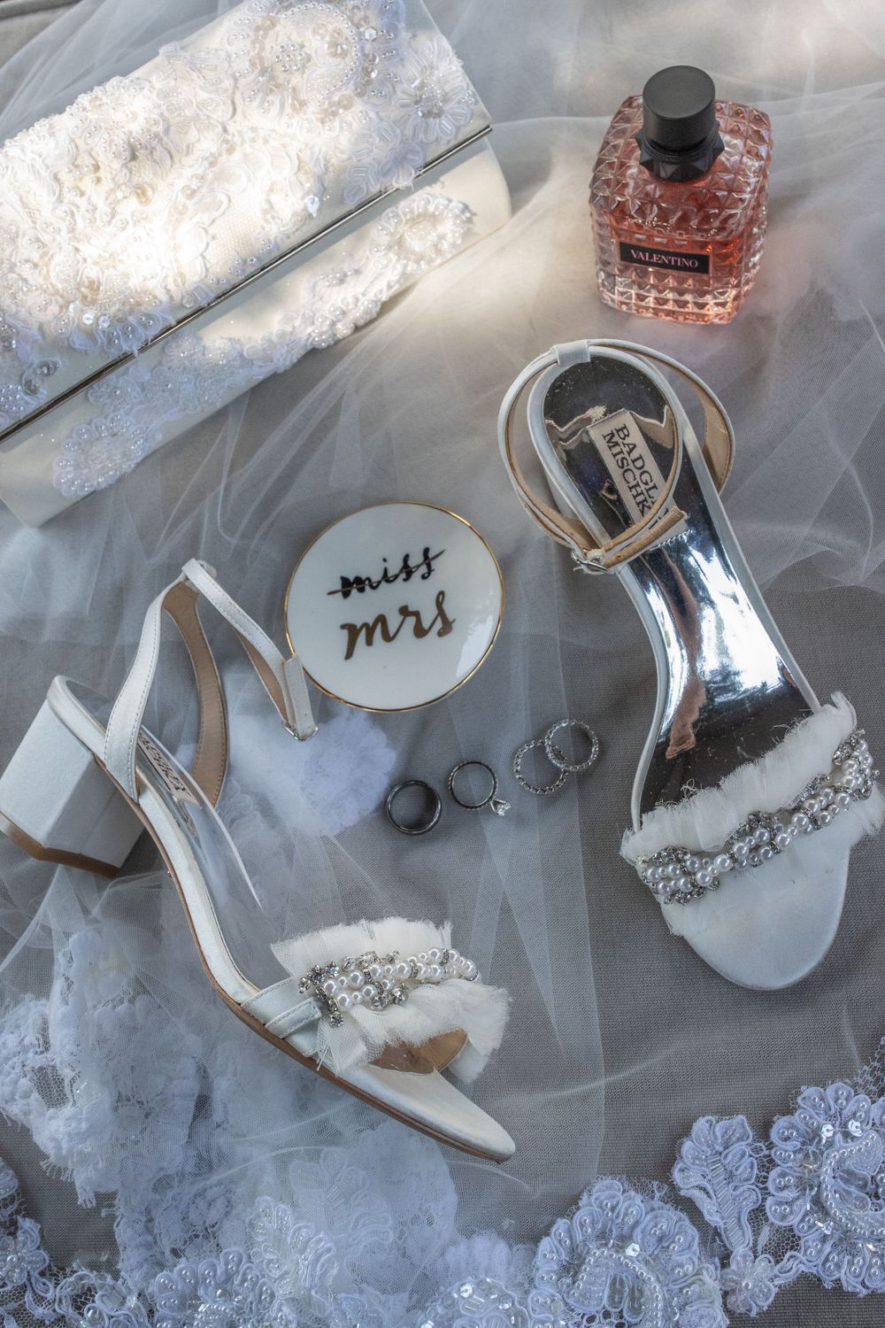 Flat lay of white bridal heels, Valentino perfume, rings, and Miss to Mrs dish on lace veil.