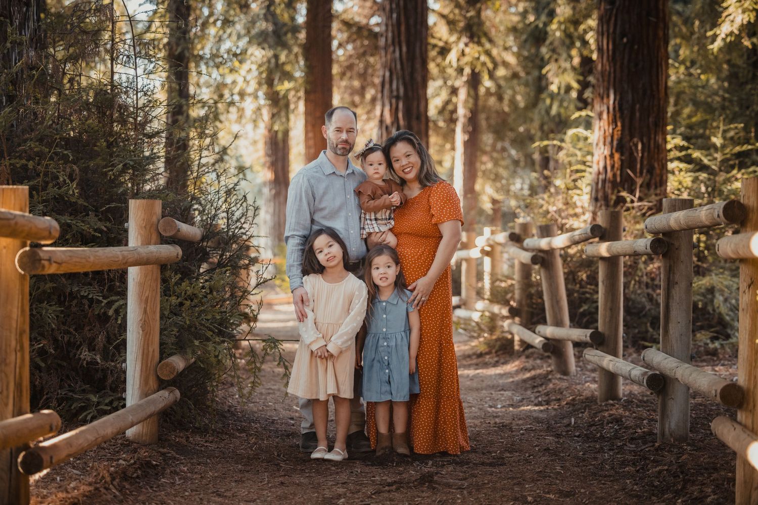 A family poses together on a rustic forest path with wooden fencing and sunlit redwood trees in the background.