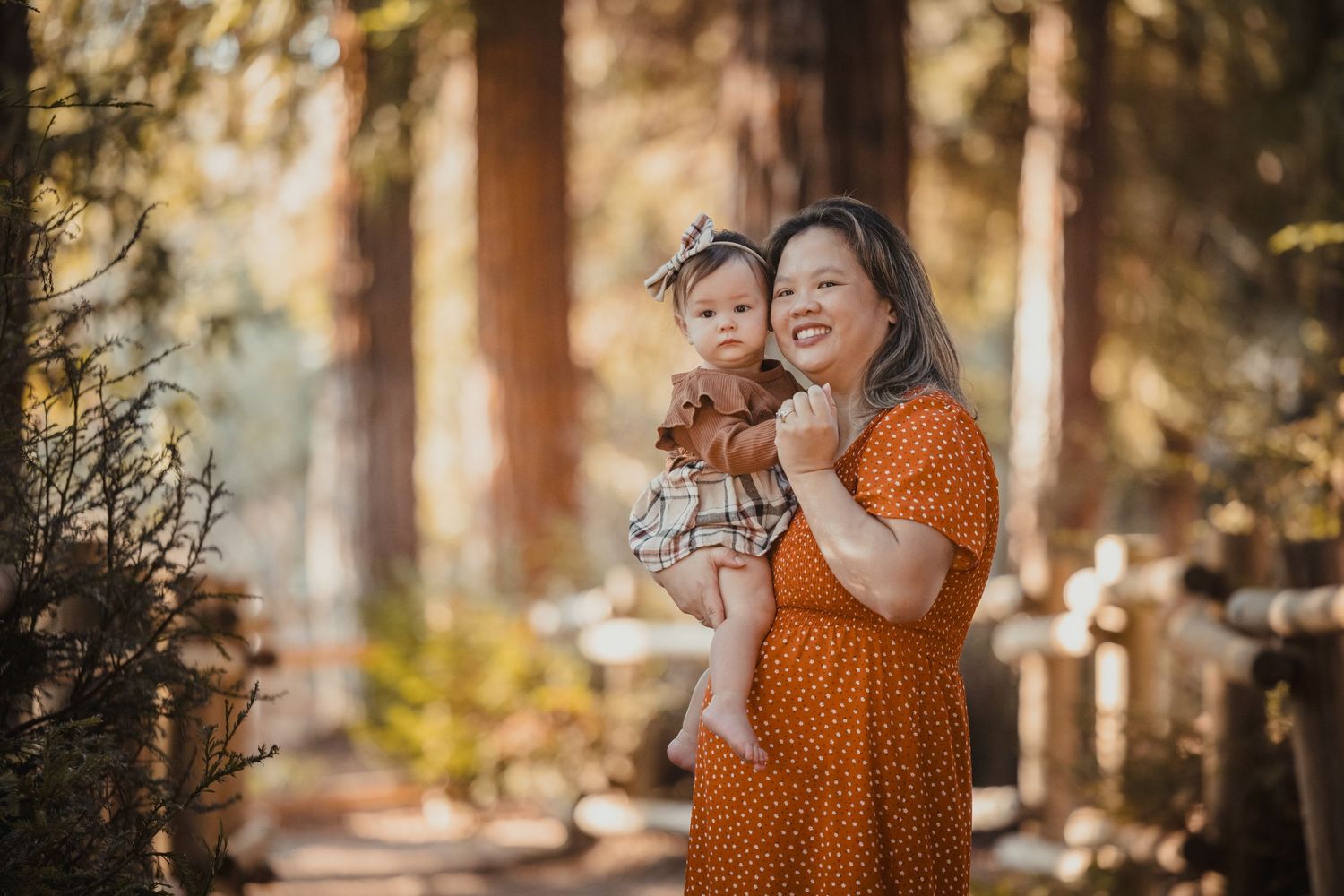 A mother and baby share a joyful moment together in a sunlit forest setting wearing orange and floral outfits.