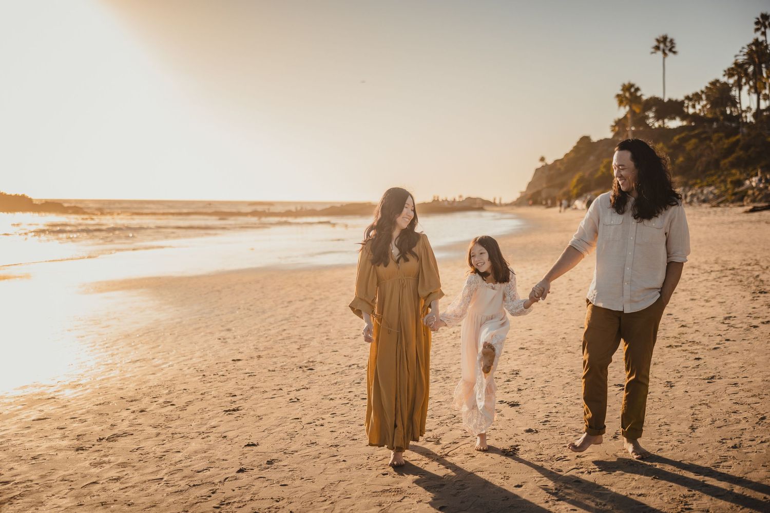 Three people holding hands while walking along a sunlit beach shore during golden hour with palm trees in the background.
