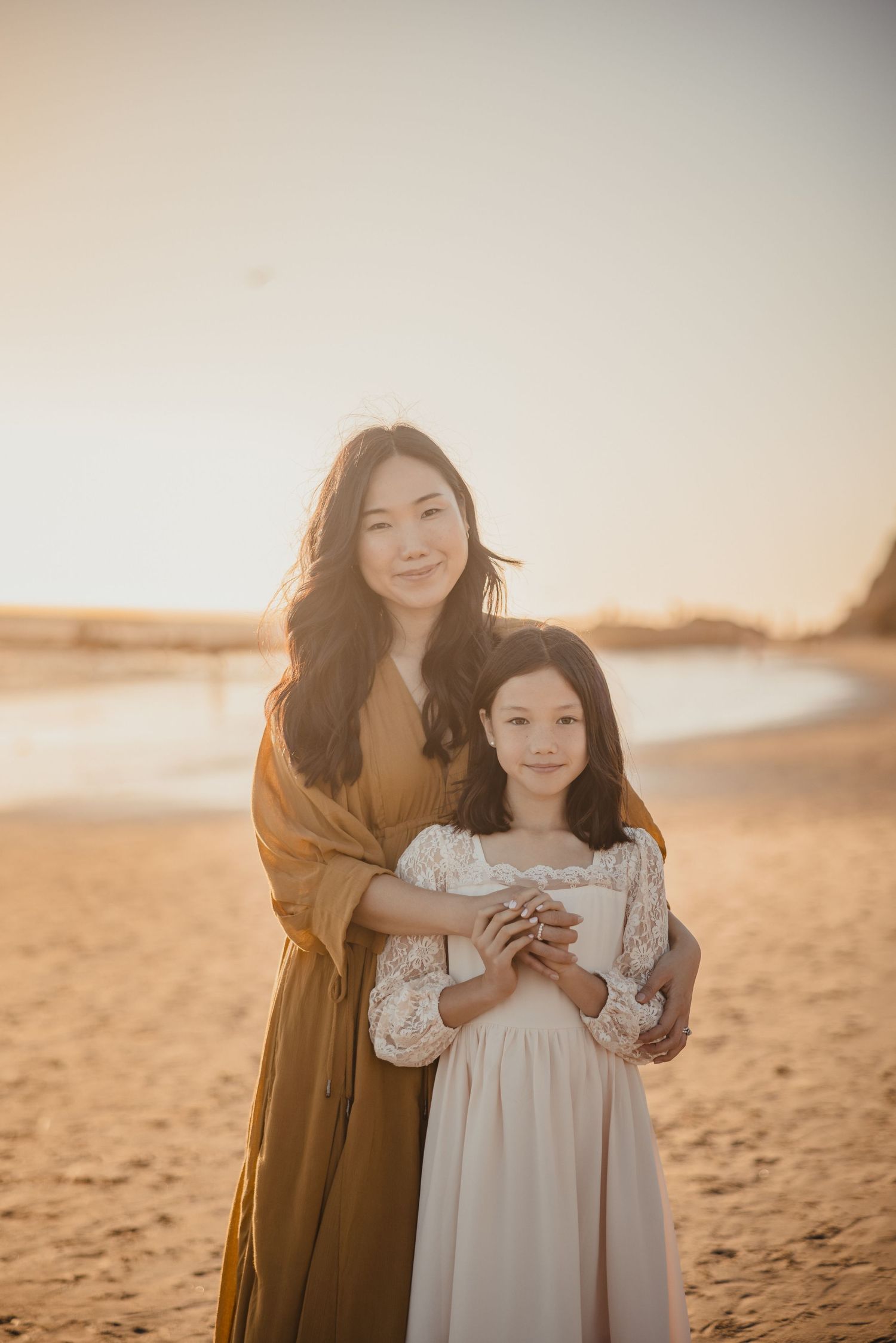 A mother and daughter stand together on a sandy beach at golden hour wearing flowy dresses in warm sunlight.