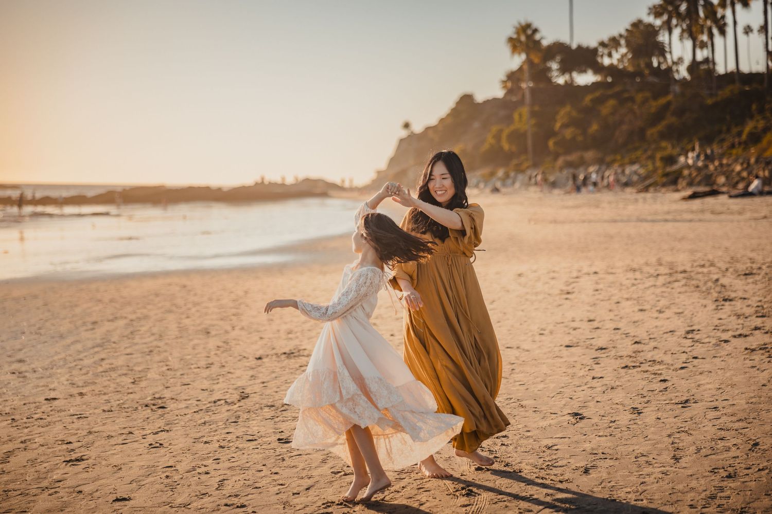 Two people in flowing dresses dance joyfully on a sunset beach with palm trees and cliffs in the background.