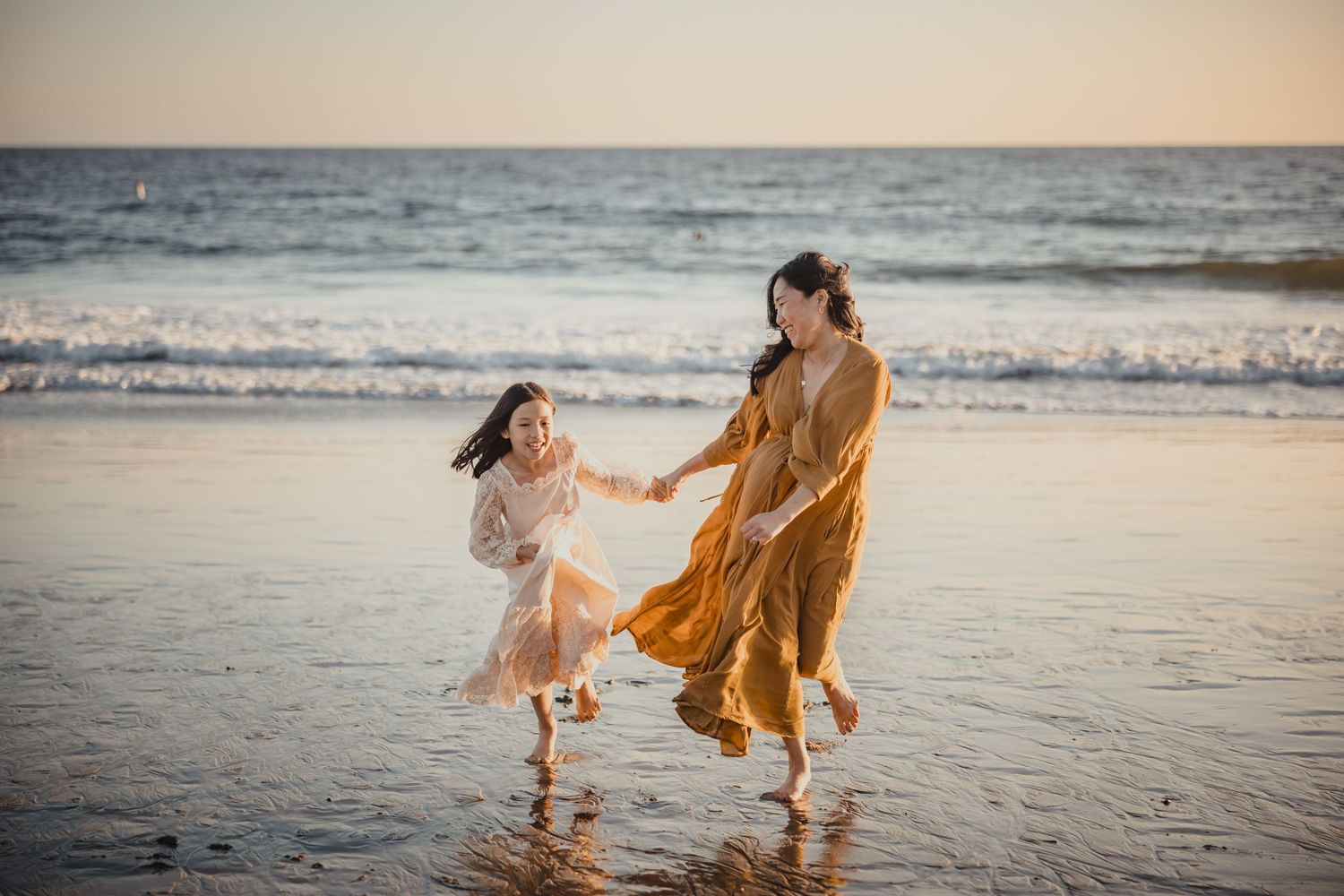 Two people in flowy dresses dance together at the ocean's edge during golden hour.