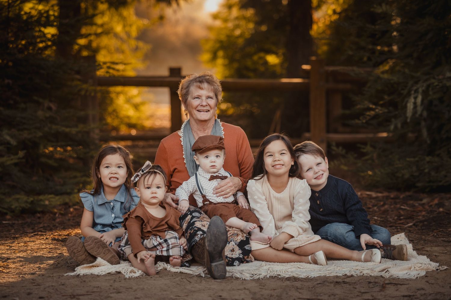 A family group sits together with their small dog on a blanket in a wooded setting during golden hour autumn light.