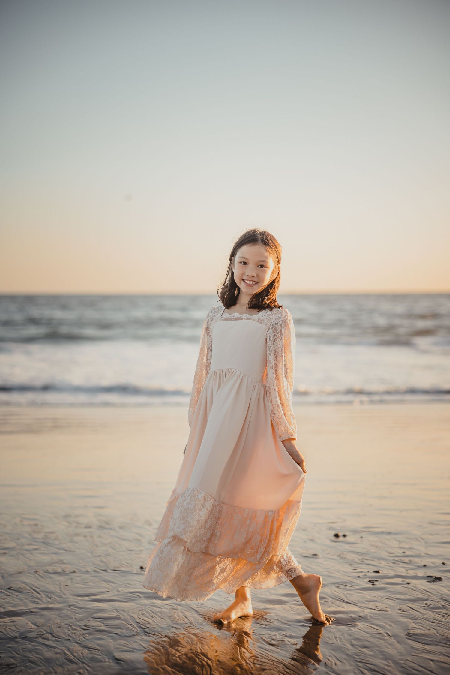 A young girl in a white lace dress splashes in ocean waves at sunset on a peaceful beach.