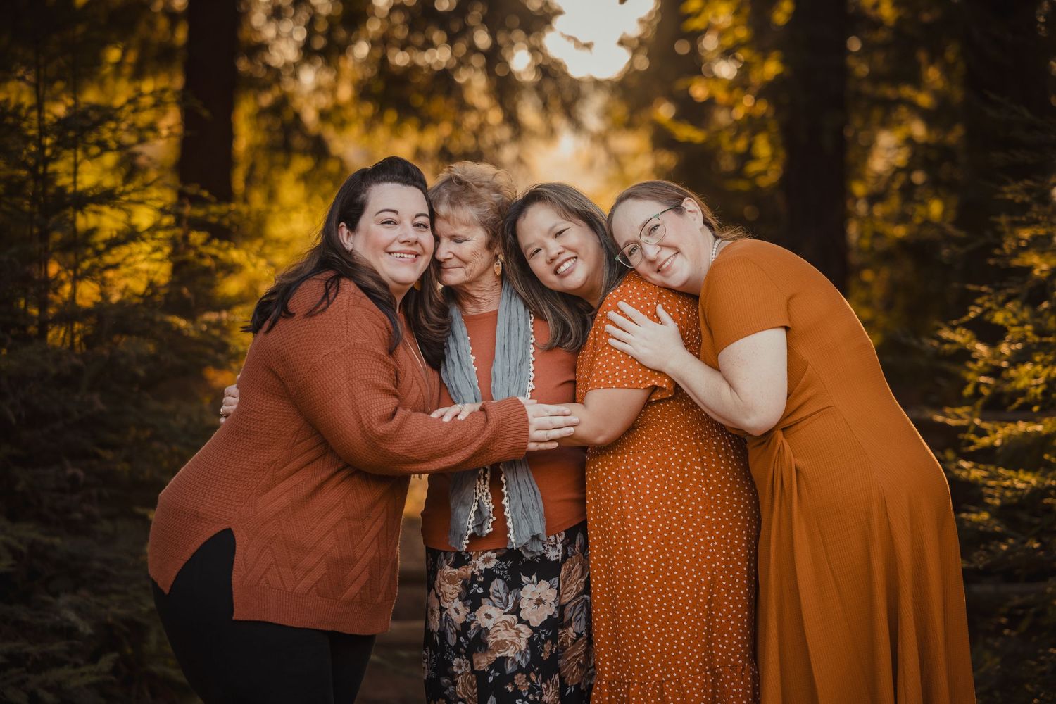 A group of four people in warm autumn tones embrace while standing in a forest with golden sunlight filtering through trees.