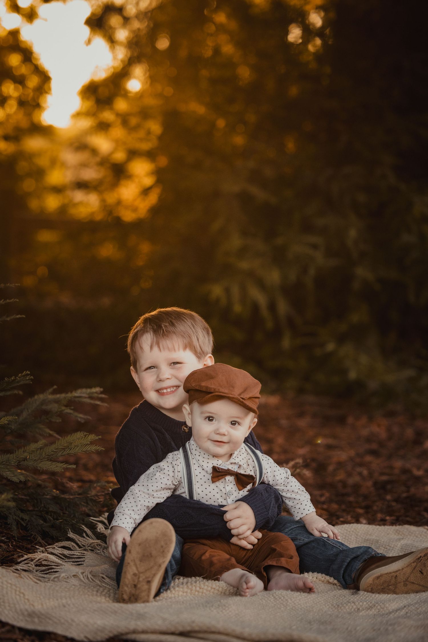 Two children sit together on a blanket in autumn forest setting with golden sunset light glowing in background.