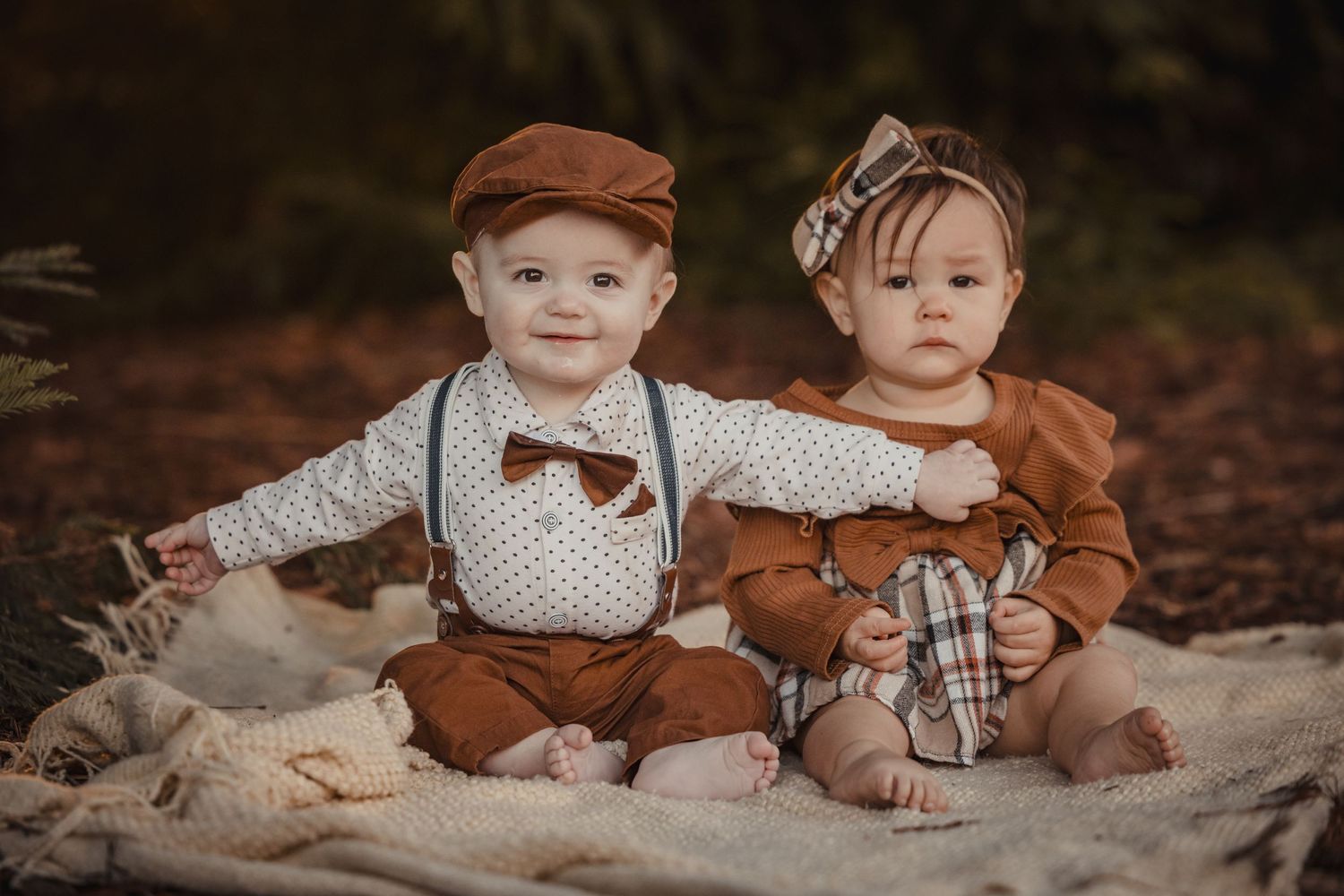 Two young children dressed in vintage-inspired autumn outfits sitting together on a blanket in a rustic outdoor setting.