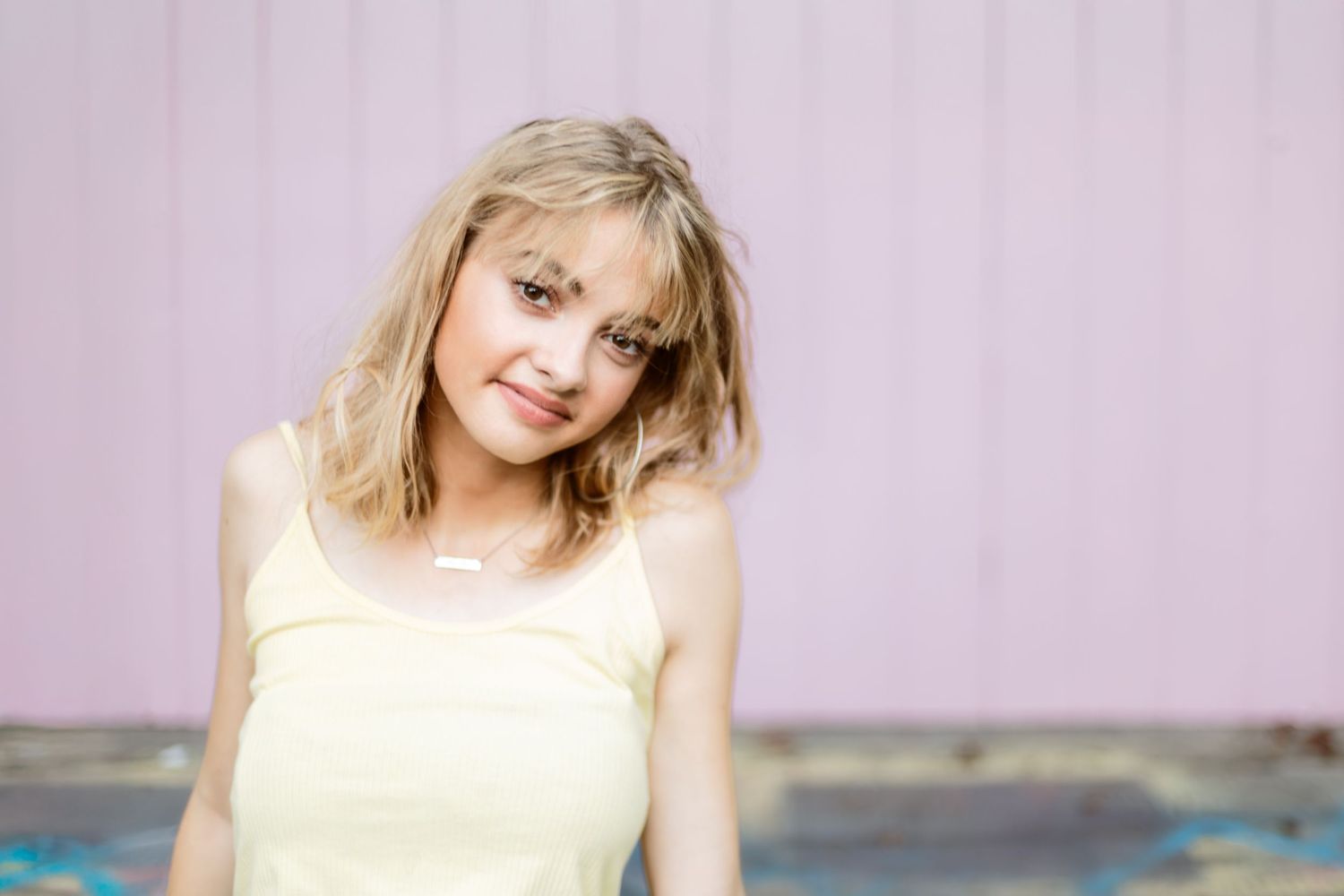 A person in a yellow tank top poses against a pink wall with tousled blonde hair.