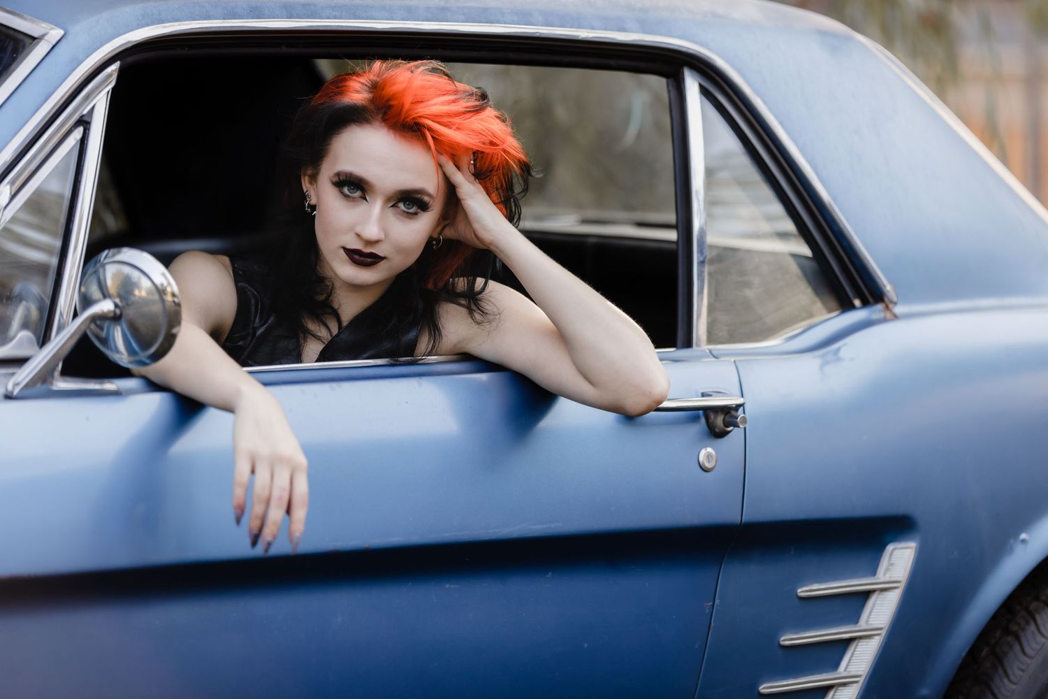 A person with bright orange hair leans out the window of a vintage blue car in an artistic portrait shot.