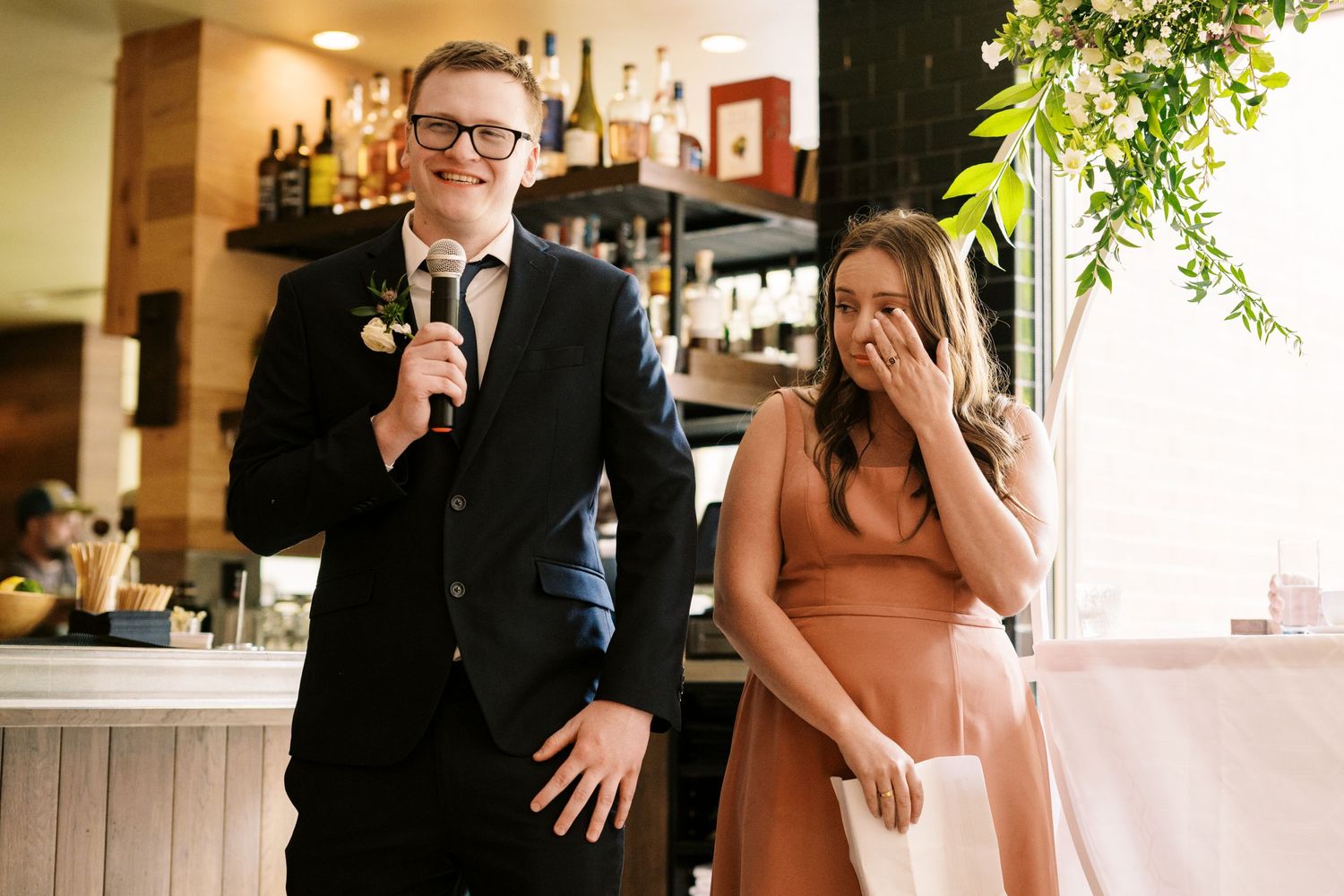 Two well-dressed people share a candid moment at a restaurant with one smiling and one covering their face in amusement.