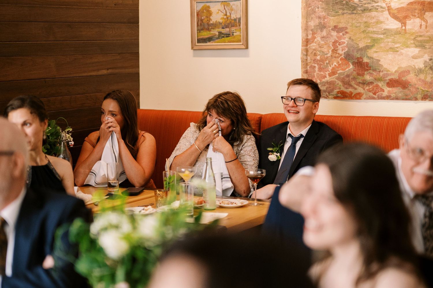 Wedding guests laugh together while seated at restaurant tables during a reception celebration.