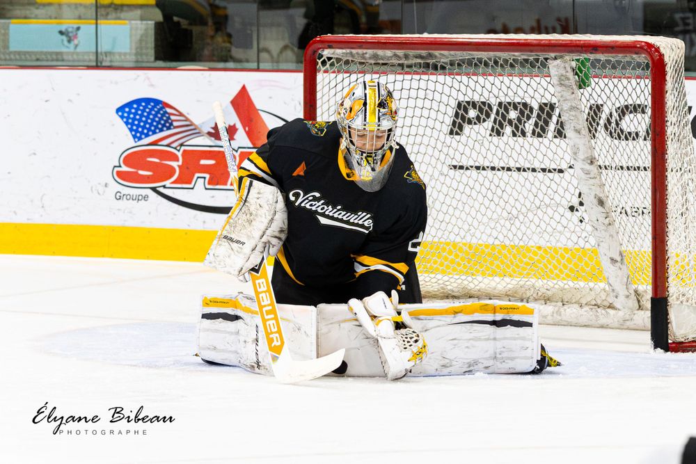 Ice hockey goalie makes a save during game with Tim Hortons advertisement visible in background.