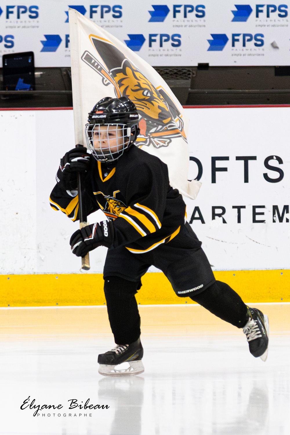 Youth hockey player in black uniform skates while carrying team flag with tiger mascot logo.