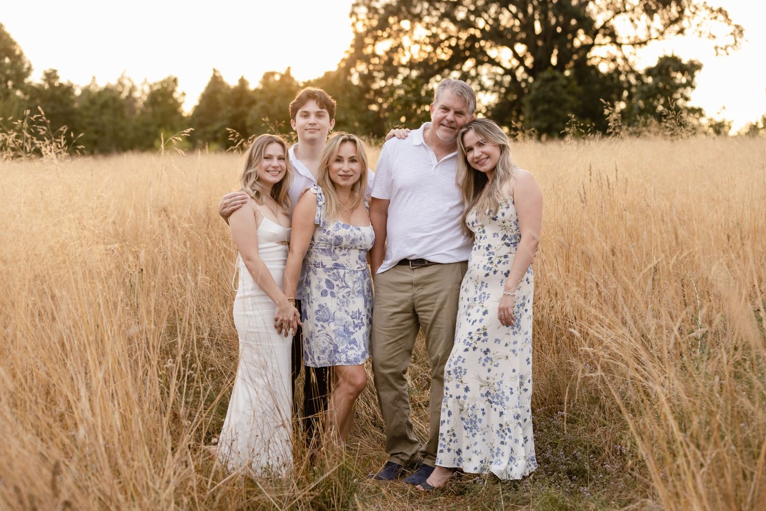 A group wearing light summer outfits poses together in a golden wheat field at sunset.