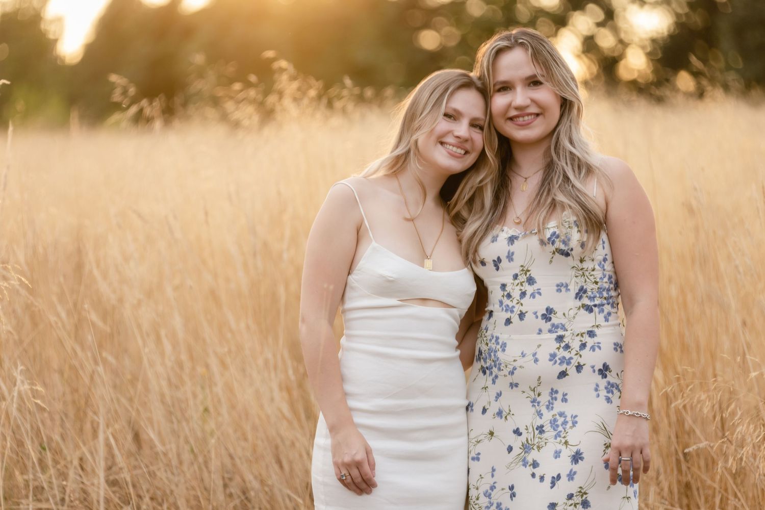 Two smiling companions in white and floral dresses stand together in a golden wheat field at sunset.