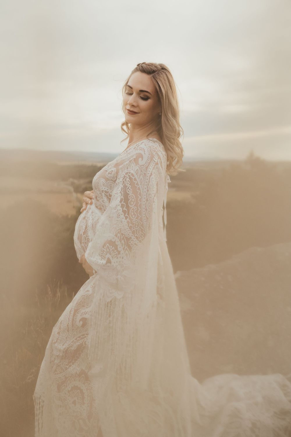 An ethereal maternity portrait features a flowing white lace dress against a dreamy outdoor backdrop.