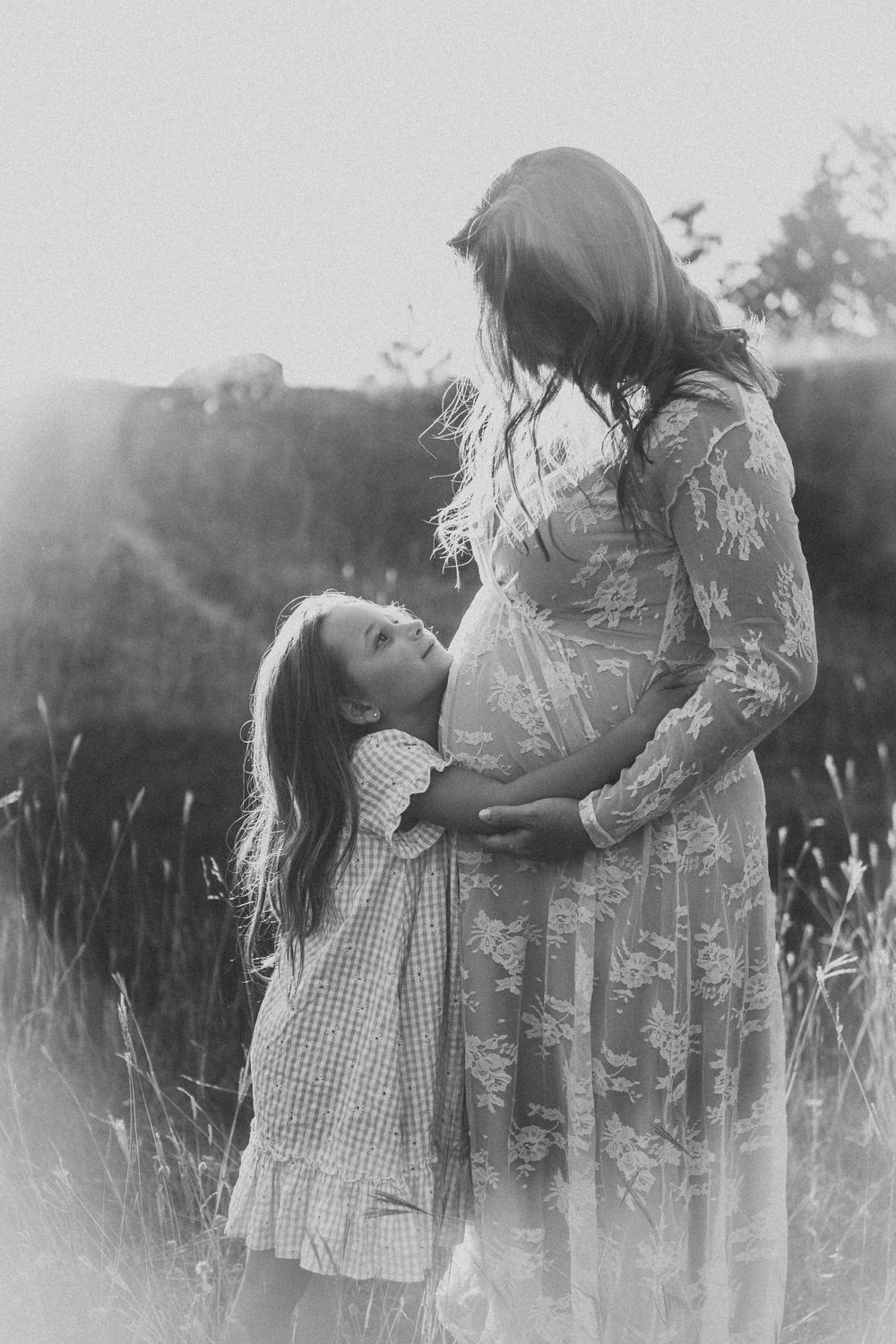 Black and white photo showing a tender family moment between expectant mother in lace dress and young child in meadow.
