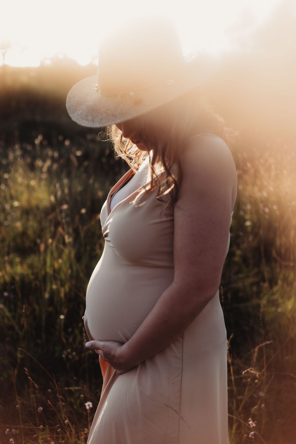 A silhouette of a pregnant figure in a white dress stands in a sunlit field at golden hour.