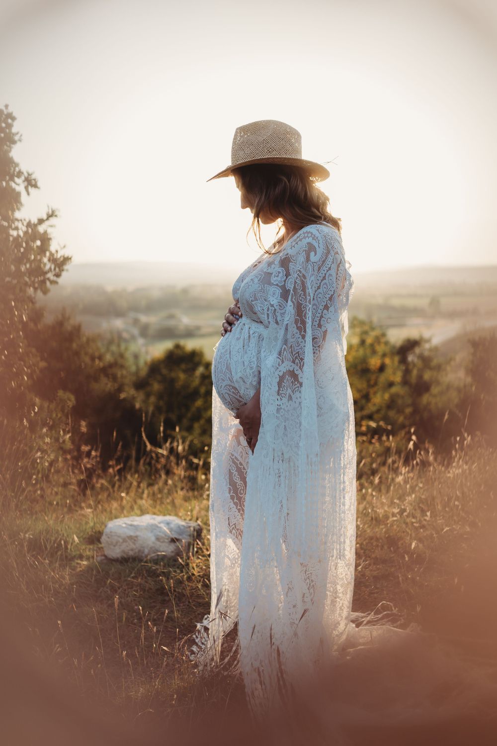 A silhouetted figure in a straw hat and flowing white dress stands on a hilltop overlook during golden hour.