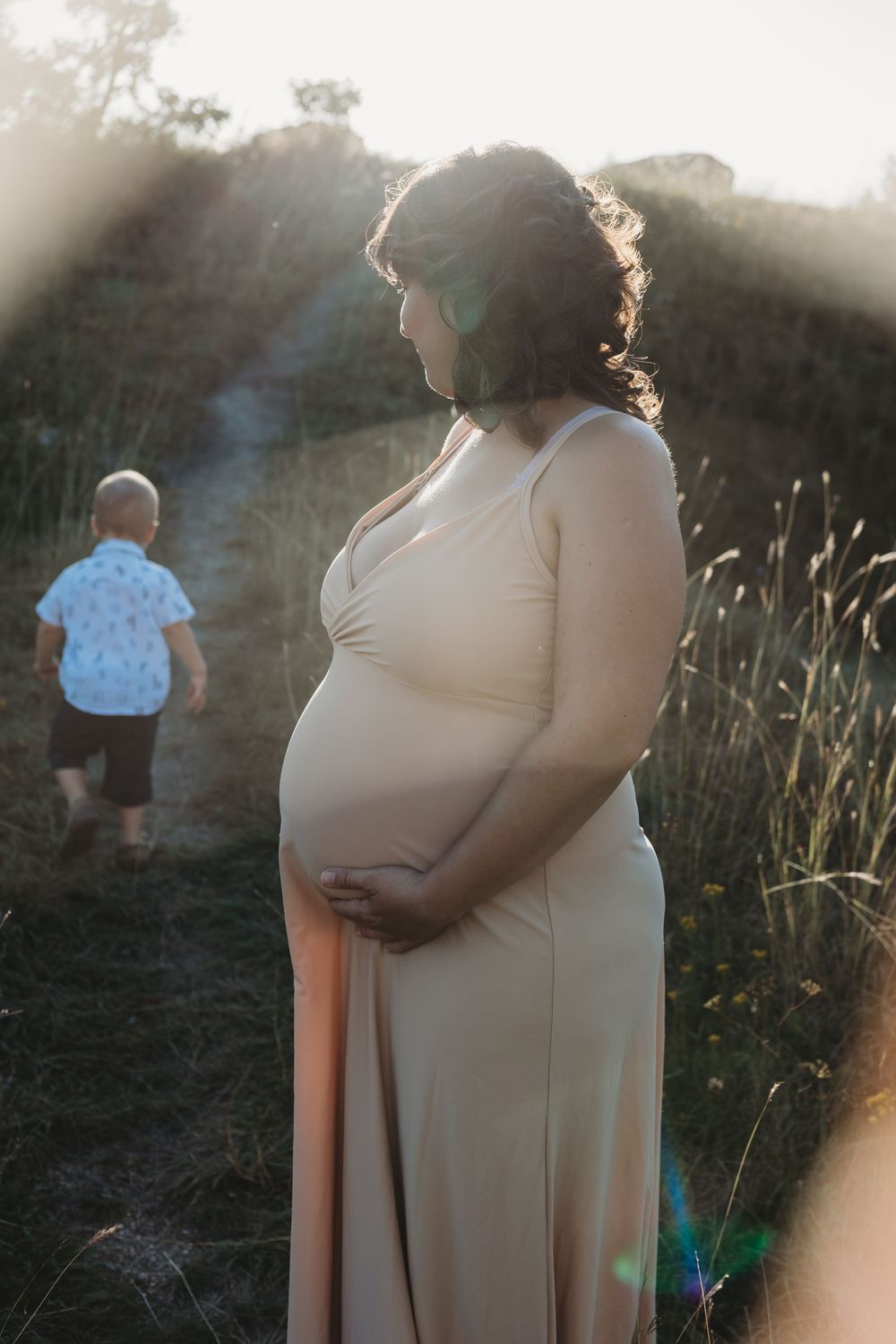 Woman in flowy beige dress stands in golden sunlight near tall grass as child plays in background.