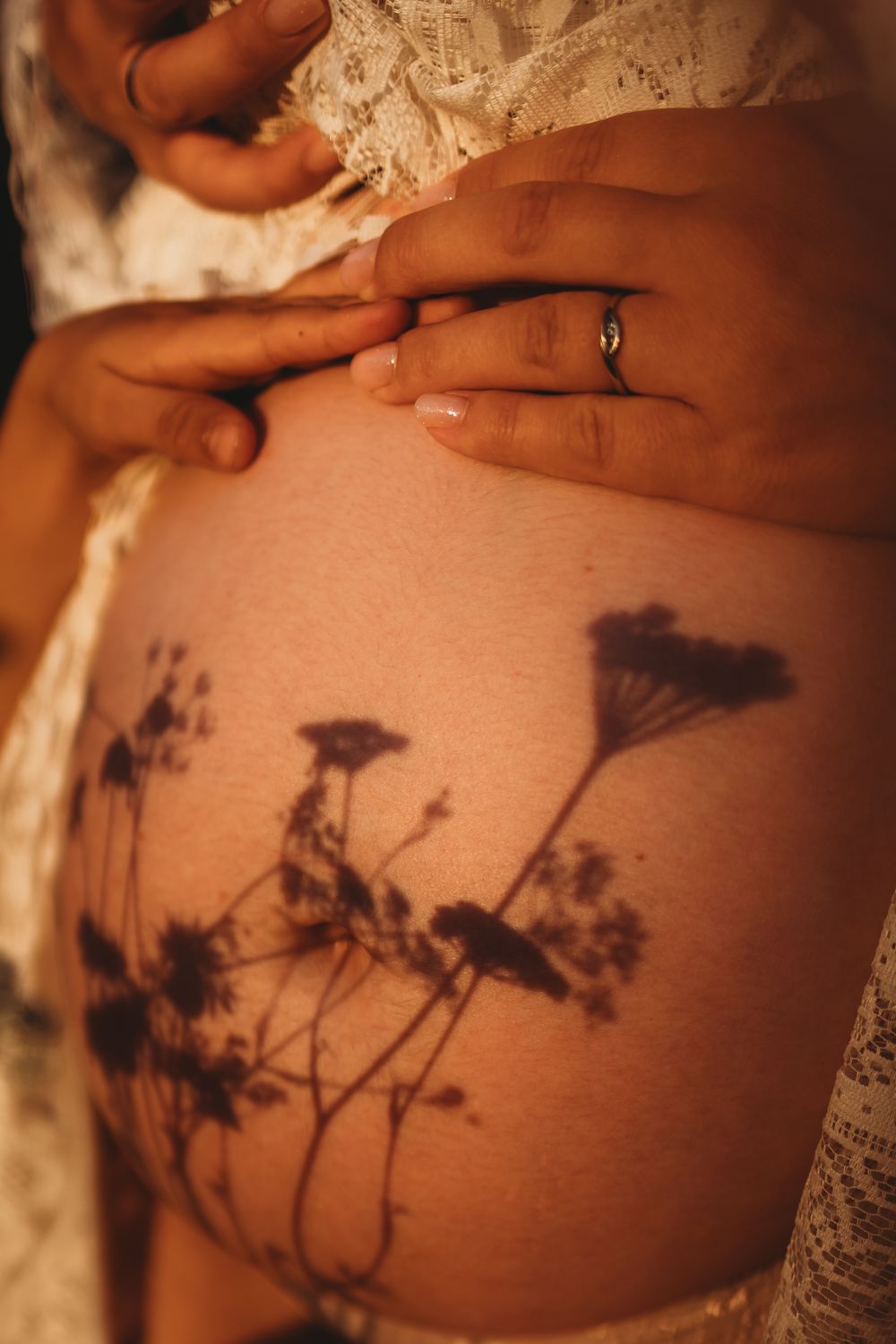 Shadows of delicate wildflowers cast on bare skin beside a hand wearing a wedding ring in warm lighting.