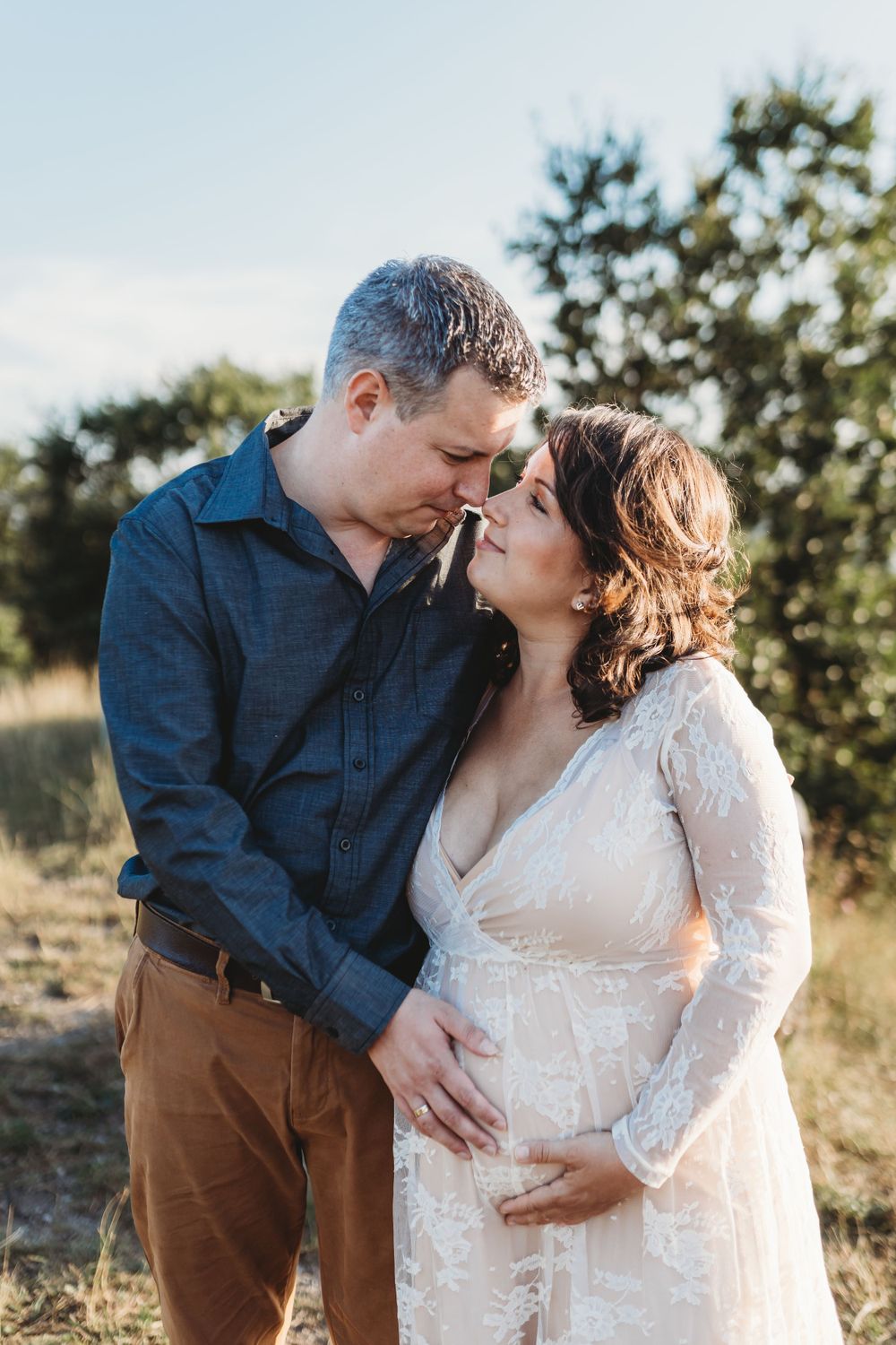 Intimate outdoor maternity photo of couple in white lace dress and blue shirt sharing tender moment.