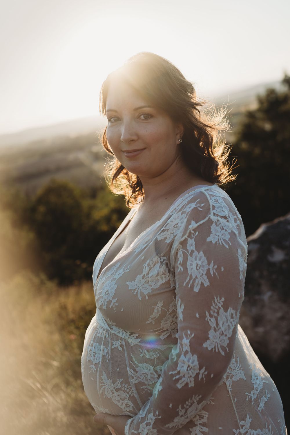 Beautiful white lace maternity dress glows in golden sunset light during outdoor photoshoot.