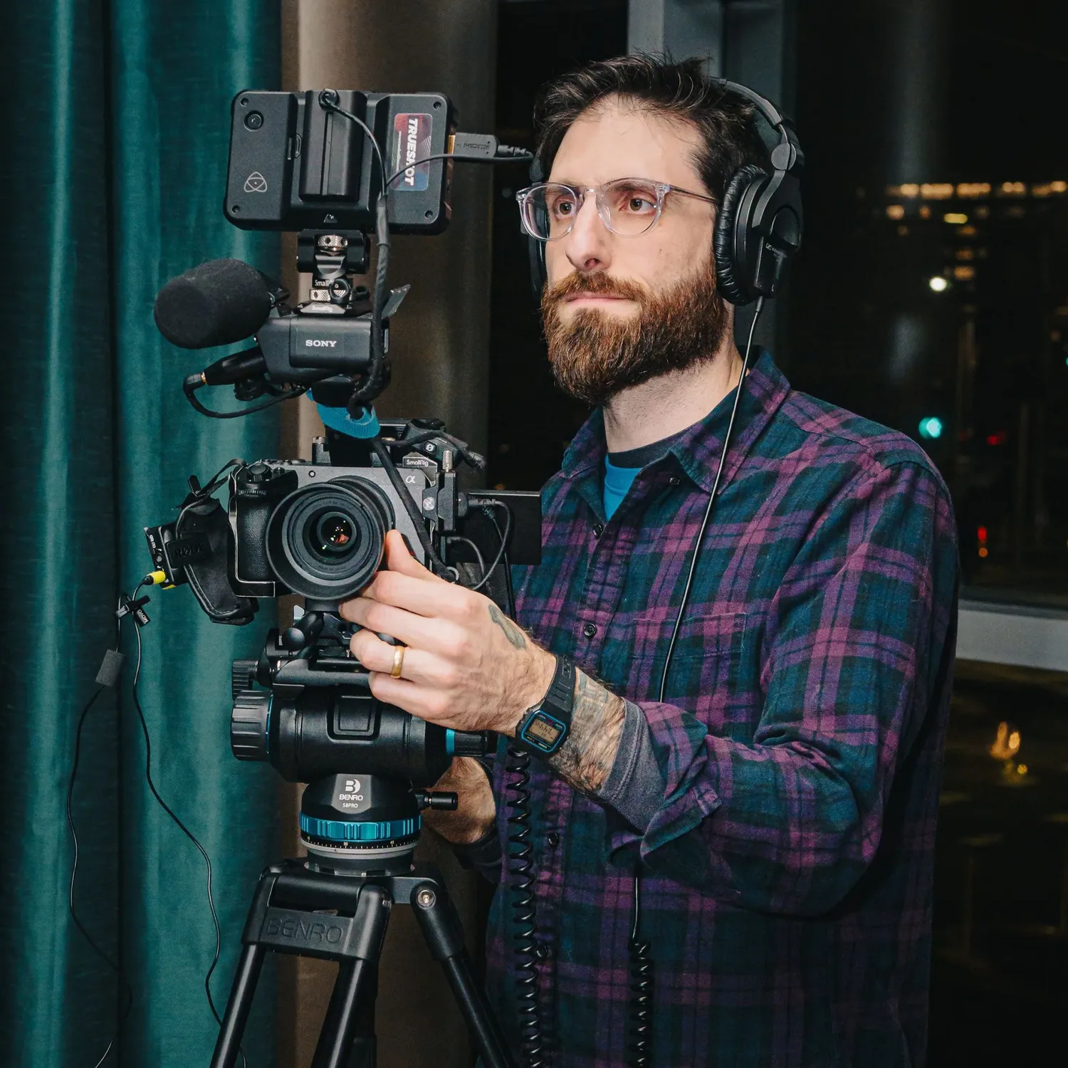 Camera operator wearing plaid shirt and headphones operates professional video equipment in dark setting with ambient lights.