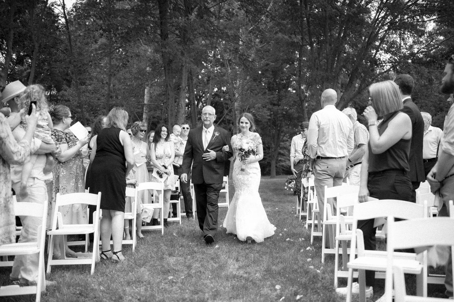 Black and white outdoor wedding ceremony in a wooded area with rows of white chairs lining a grassy aisle.