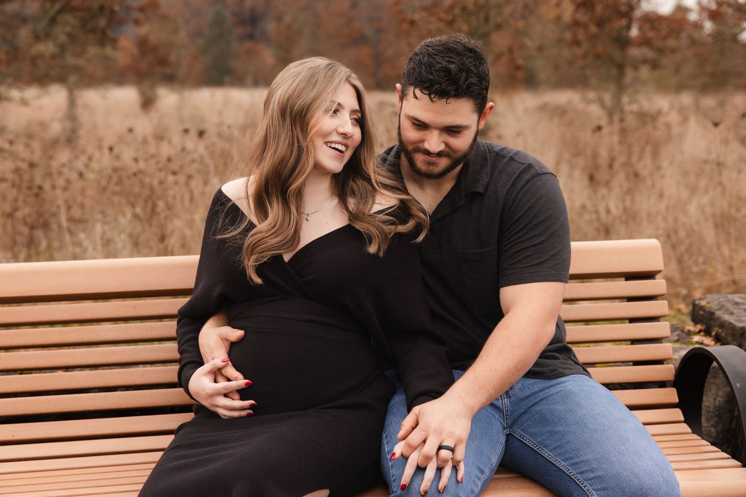 A couple sits together on a wooden bench during an autumn maternity photoshoot in a park.