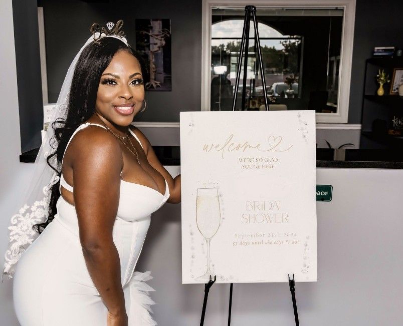 A bride in a white wedding dress stands next to a welcome sign board at her reception venue.