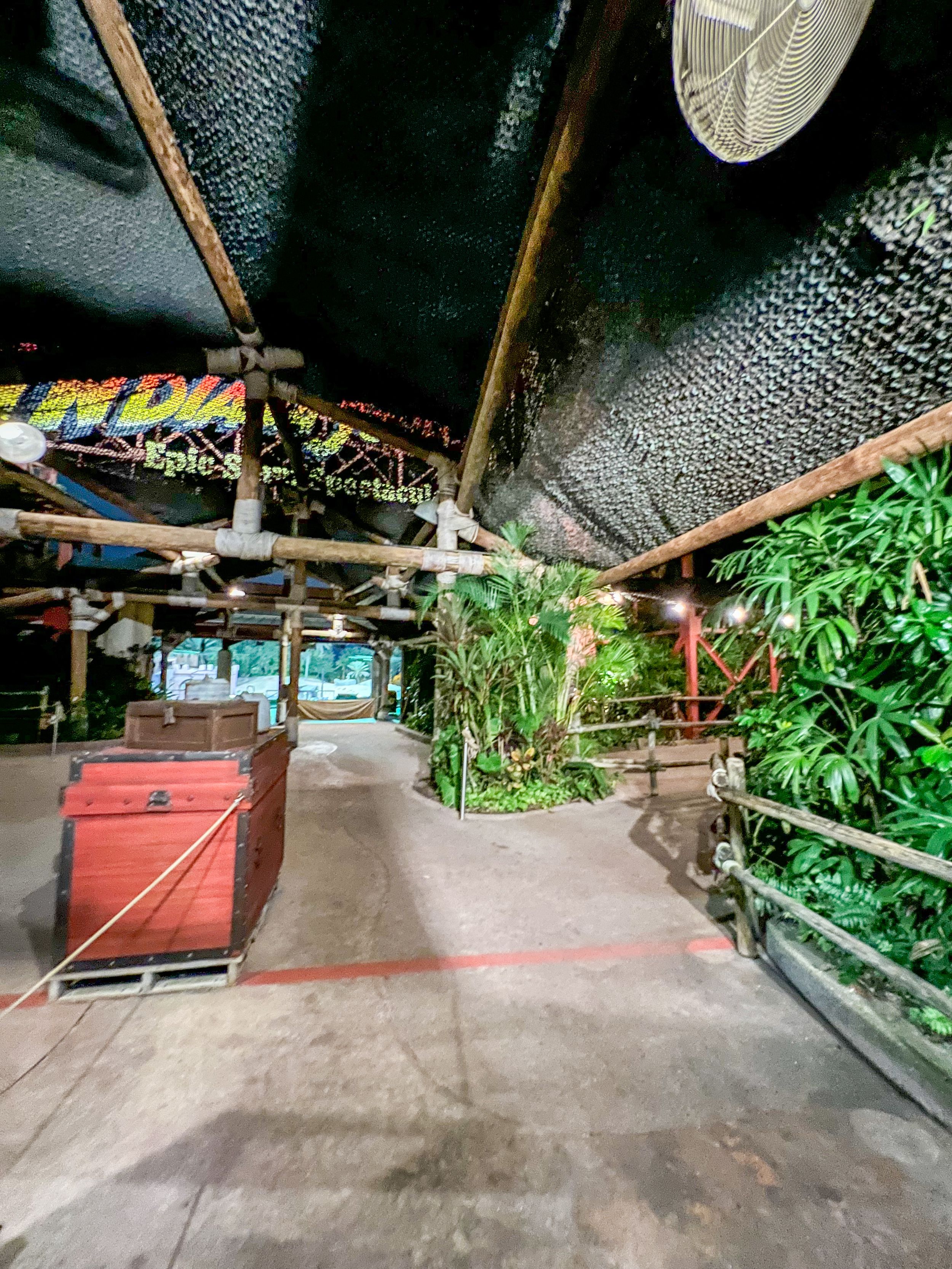 Covered walkway with tropical plants and a red container in a dimly lit open-air structure.