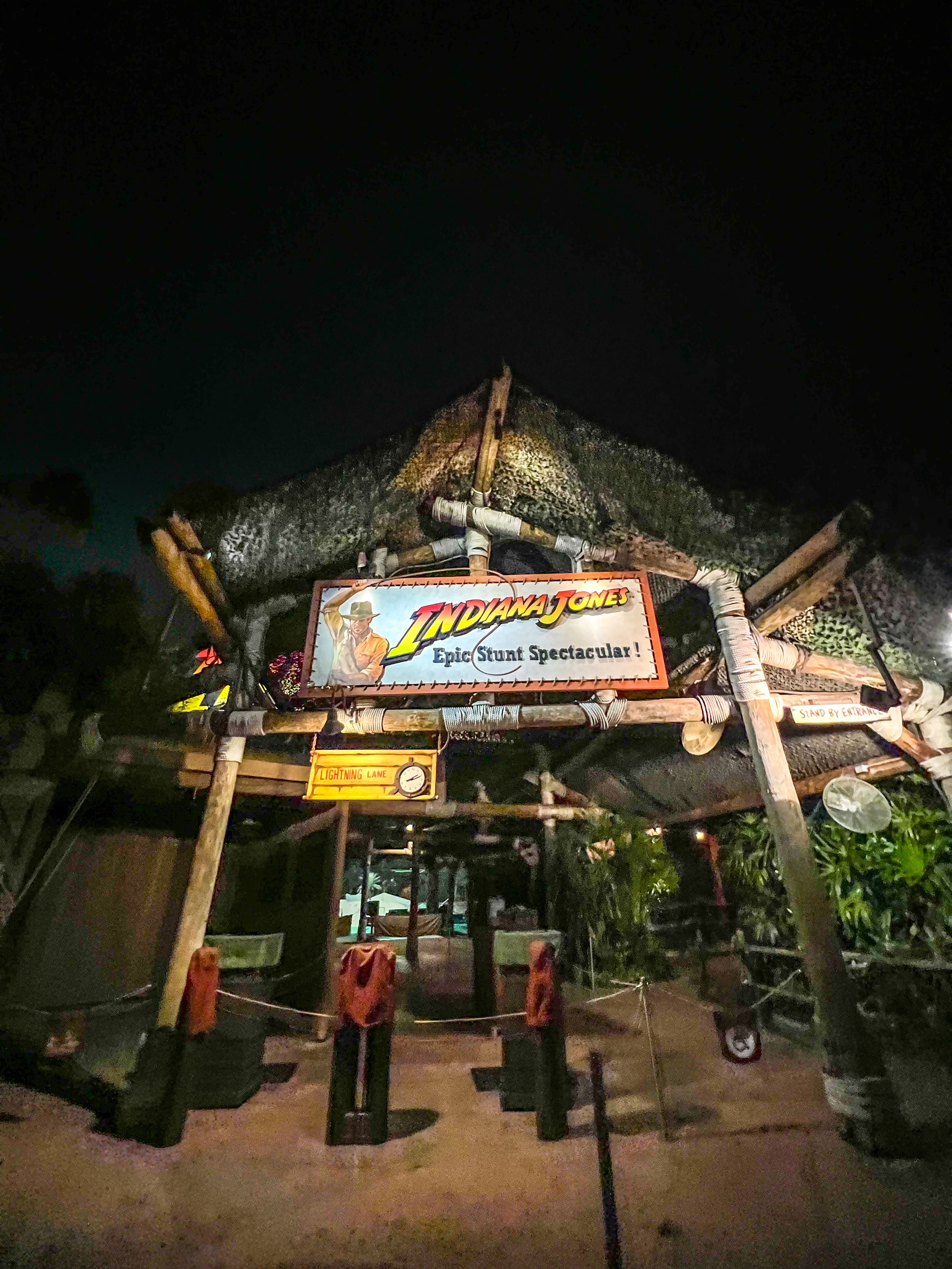 Illuminated tiki-style entrance sign for a tropical beach bar at night, showcasing rustic wooden poles and palm trees.